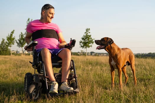 Homosexual man in a dark wheelchair with a back injury enjoys playful moments with his pet dog in a sunny meadow.