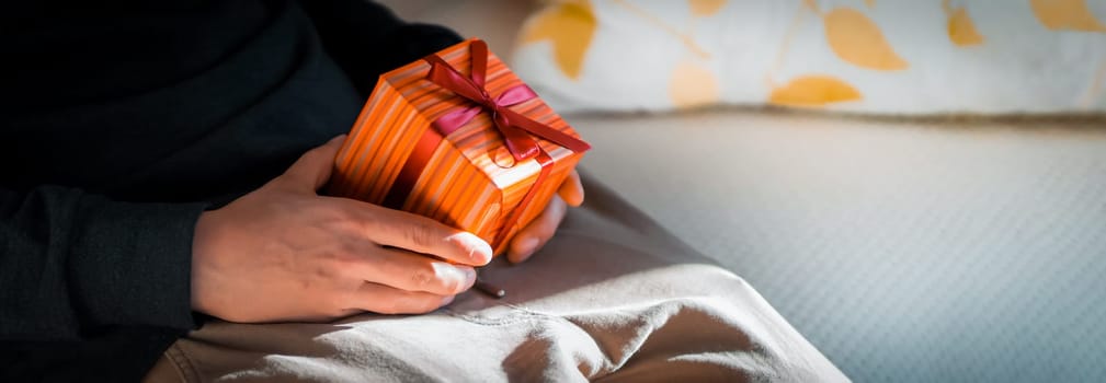 A funny erotic gift for a partner for a holiday, anniversary, Valentine's day, a young man sits on the couch and holds a small gift wrapped in a box, tied with a ribbon in an intimate place.