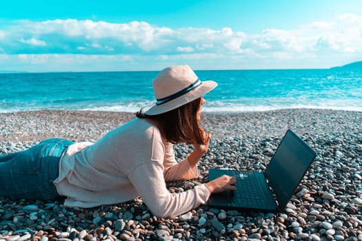 A young girl in a light hat and casual sweater lies on the beach by the sea with a laptop on a sunny day, works, studies, buys tickets during trip, a woman rests on vacation and types on the keyboard.