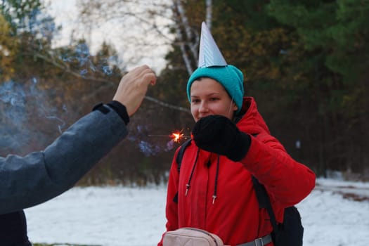 Two cheerful friends embrace the winter chill, wearing colorful hats and laughing as they ignite sparklers in a snowy forest setting
