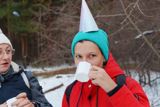 Two friends enjoy sweet snacks and warm beverages while wearing colorful party hats in a serene snowy forest.