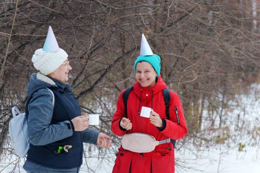 Two friends in festive party hats enjoy warm beverages while standing in a snowy area.