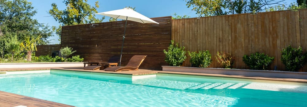 Relaxing backyard swimming pool with turquoise water, wooden deck, lounge chairs, umbrella, and green garden