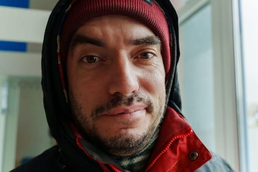 A close-up of a man wearing a red beanie and jacket, standing indoors during a chilly winter afternoon in an urban setting