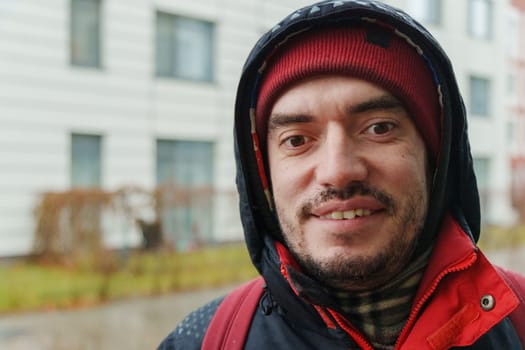 A man braves the chill outdoors, wearing a red beanie and a coat, showcasing a playful grin against a city backdrop