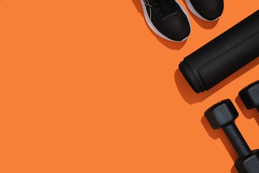 Black workout equipment, dumbbells, mat, sneakers on orange background. Fitness, sports items, healthy lifestyle concept. Space for your text