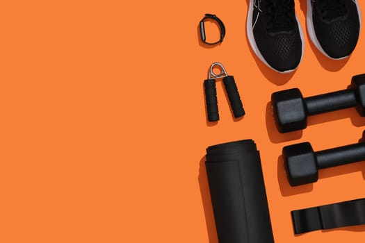 Black exercise equipment, dumbbells, mat, elastic band, fitness band, fitness band, sneakers on orange background. Fitness, sports items, healthy lifestyle. Place for your text