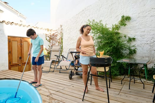 A serene backyard scene featuring a woman preparing a barbecue while a boy cleans the pool.