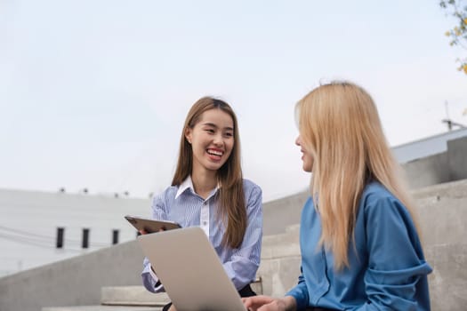 Two businesswomen engaged in a discussion outdoors, sharing ideas while using a laptop and a tablet in a collaborative environment.