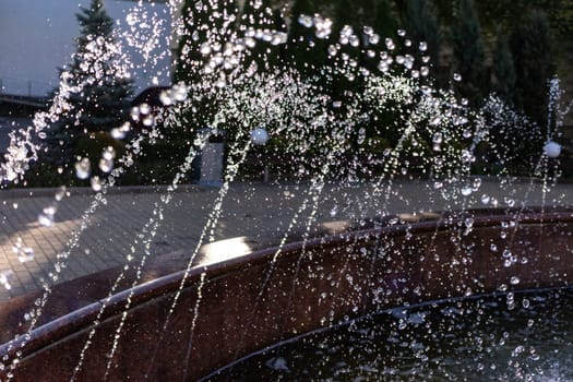 Water streams from a fountain in a city park, creating sparkling droplets that shimmer in the evening light. Surrounding greenery adds a serene atmosphere to the scene.