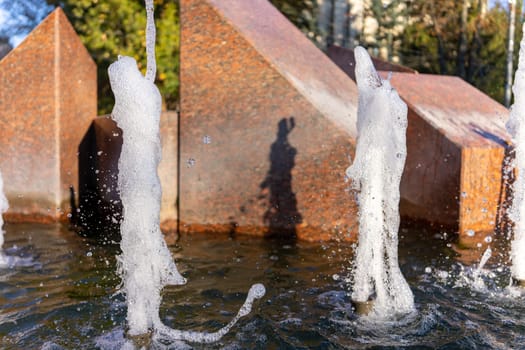 Vivid water jets rise and fall in a modern urban park. Sunlight glimmers on the droplets, casting shadows on the textured backdrop, showcasing a playful interaction of light and water.