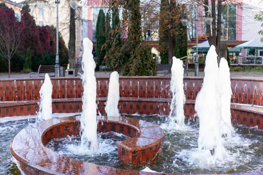 Multiple water fountains with dancing streams enhance the beauty of a city park in autumn. Lush trees surround the area, creating a tranquil and inviting environment for visitors.