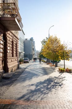 A peaceful urban walkway features colorful autumn leaves under clear blue skies. Pedestrians stroll along the path beside charming buildings, creating a serene morning atmosphere. Ukraine, Vinnytsia, 2021.01.15