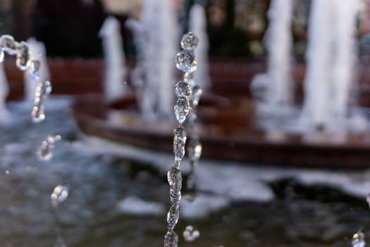 Water droplets emerge from a fountain, creating a mesmerizing display as they fall into the basin below. Sunlight sparkles on the water, enhancing the serene atmosphere of the park.