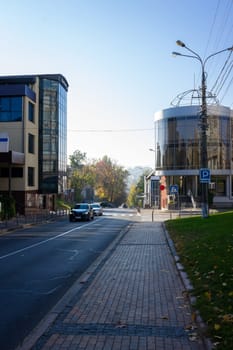 A peaceful urban street in autumn showcases modern glass buildings. A car drives by on a quiet road lined with trees and colorful leaves under a clear blue sky. Ukraine, Vinnytsia, 2021.01.15