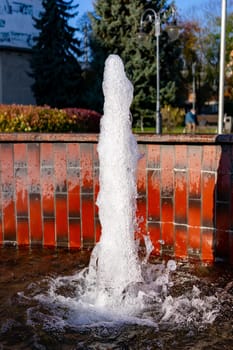 A water fountain erupts into the air in a lively city park. Sunlight glimmers on the water, surrounded by greenery and brickwork. People enjoy the serene atmosphere nearby.