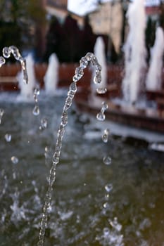 Water droplets arc gracefully from various fountains, creating a captivating display as the sun sets behind lush trees in a vibrant city park.