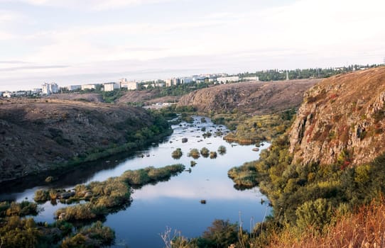 A huge wide river flows between steep banks. A city is visible on the horizon. Autumn forest on the banks of the river. Cliffs of granite stones. Bushes grow in the middle of the river. southern bug