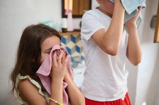 Two children are seen wiping their faces with towels after washing, embodying morning hygiene routines and freshness. Smiles convey joy and a healthy lifestyle in a home bathroom environment.