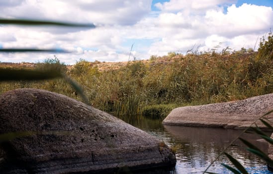 Beautiful blue lake with boulder figures. There is a lake in a mountain valley, reeds grow around it. Fantastic granite gray boulders protrude from the water, close-up. Aktovsky Canyon, Ukraine.