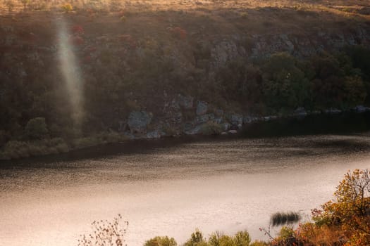 a huge wide river flows among steep banks. autumn forest on the banks of the river. cliffs of granite stones. a ray of sunlight falls on the water surface. southern bug, ukraine
