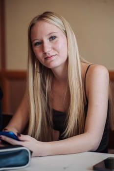 Blonde high school student sitting at a cafeteria table, engaged with her smartphone, connecting with friends during lunch break