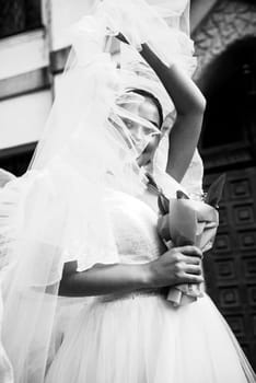 Bride in a white dress with a veil covering her face. The girl holds a bouquet of lilies of the valley in her hands, she looks at the camera through the veil. Stylish hairstyle. black and white photo