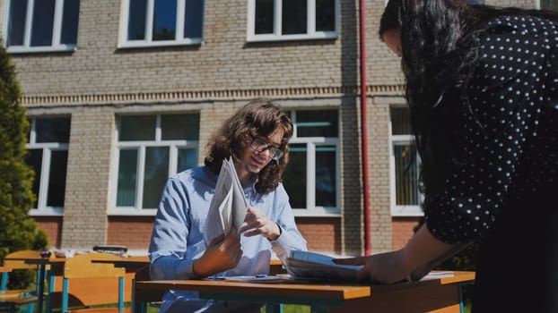 Two high school students are working together on a project at a table outside their school building on a sunny day