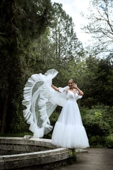 beautiful bride with long veil against the background of trees and old fountain. brunette girl in long white dress, she has smooth hair, old ruined fountain behind. lush veil flutters in wind