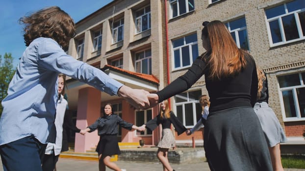 Group of cheerful students holding hands and playing in front of their school on a sunny day