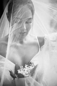 close-up of a bride in a white dress with a veil covering her face. the girl is holding a bouquet of lilies of the valley. she has a stylish hairstyle, her hair is styled in a bun. black white photo
