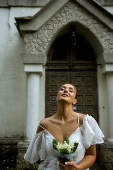 Bride in a white dress and with a bouquet of lilies of the valley. Dress with open shoulders. Behind is an old church of the 18th century. The girl has a stylish hairstyle, her hair is put in a bun.