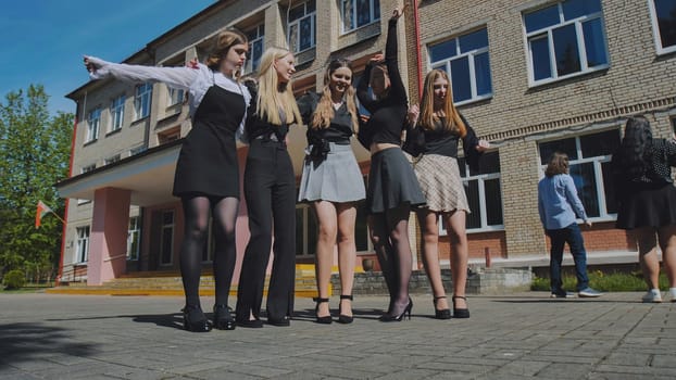 Five teenage girls happily posing in front of their high school building on a sunny day, marking the end of the school year
