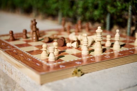 A close-up outdoor photograph featuring a wooden chessboard with scattered chess pieces.