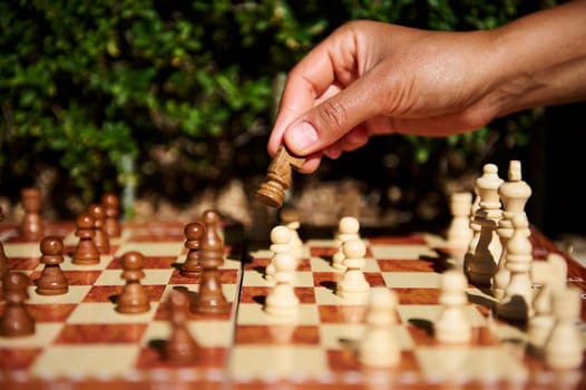 A hand holding a chess piece during a game on a wooden chessboard outdoors, promoting mental strategy and competition.