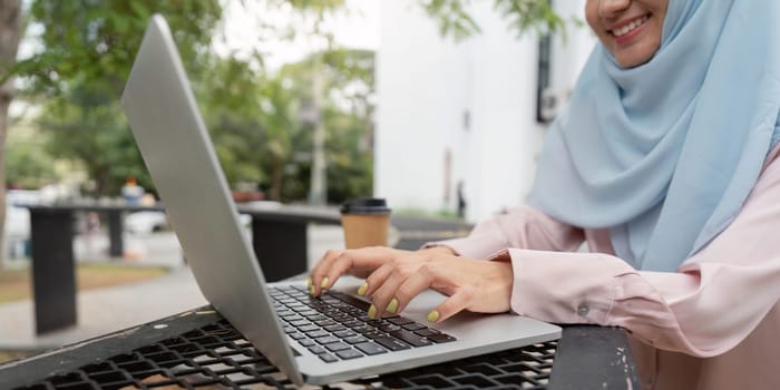 A focused Muslim woman in a hijab types on her laptop, with a coffee cup nearby, illustrating a modern approach to work and productivity in a serene outdoor setting.