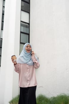 A joyful Muslim woman in a hijab engages in a phone conversation while holding a coffee cup, reflecting a dynamic and professional lifestyle in a modern business setting.