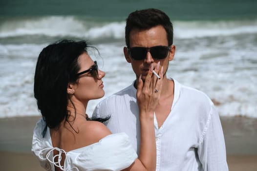 stylish young beautiful couple in love smokes a cigarette on the beach by the sea in summer