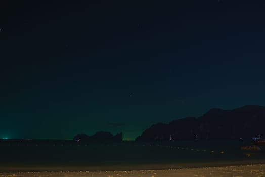 A serene beach at night with a sandy shoreline, calm sea, starry sky, and silhouetted limestone cliffs in Thailand's Phuket or Koh Phi Phi islands.