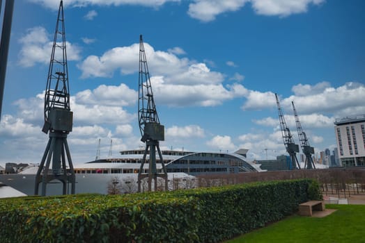Iconic cranes at Royal Victoria Dock in London stand against a blue sky. A hedge and bench in the foreground contrast with a modern building in the background.