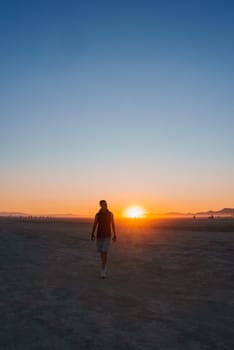 A person walks towards the camera in a desert at sunset, silhouetted against orange and blue skies. Groups of people and low mountains are visible in the distance.