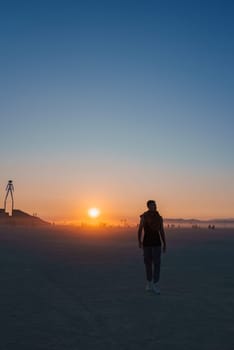 A tall wooden figure stands silhouetted against a vibrant sunset in Black Rock Desert, Nevada. Scattered participants walk across the flat, dusty ground.