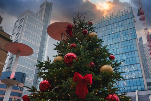 A Christmas tree with red and gold ornaments stands before modern skyscrapers in London. Mushroom shaped structures and a crane highlight urban development.