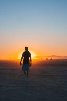 A person walks silhouetted against a vibrant orange and blue sunset sky in a desert. Distant figures on bicycles enhance the festival atmosphere.