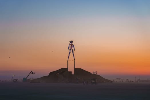 A large, light adorned human sculpture stands on a mound in Black Rock Desert. The sky's warm gradient and distant structures enhance the festival scene.