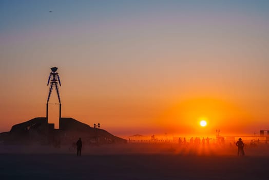 The iconic Burning Man effigy stands silhouetted against a warm sunset in the Black Rock Desert, Nevada, with participants visible in the distance.