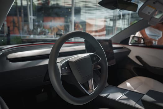 The image captures a modern car interior with a sleek steering wheel and large touchscreen. Through the windows, blurred glass structures and greenery suggest an urban setting.