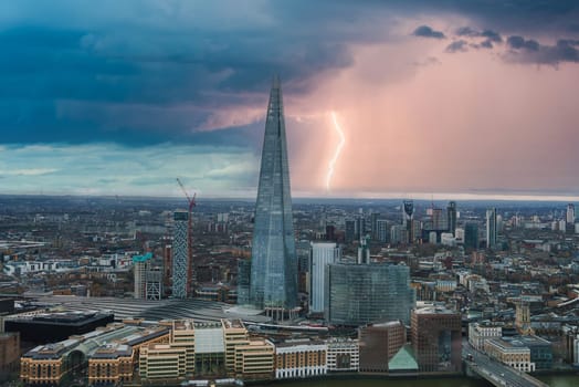 The Shard stands prominently in London's skyline as a bolt of lightning illuminates the stormy sky. The Thames River and diverse architecture are visible.