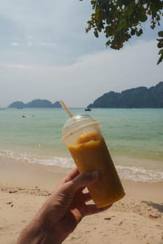 A vibrant yellow smoothie in a plastic cup held against a serene beach backdrop, with turquoise waters, lush islands, swimmers, and boats in Thailand.