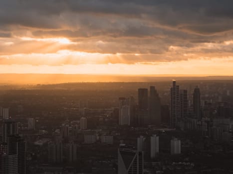 The image shows London's skyline at sunset, with skyscrapers silhouetted against a vibrant sky. Sun rays pierce through clouds, casting a golden hue.
